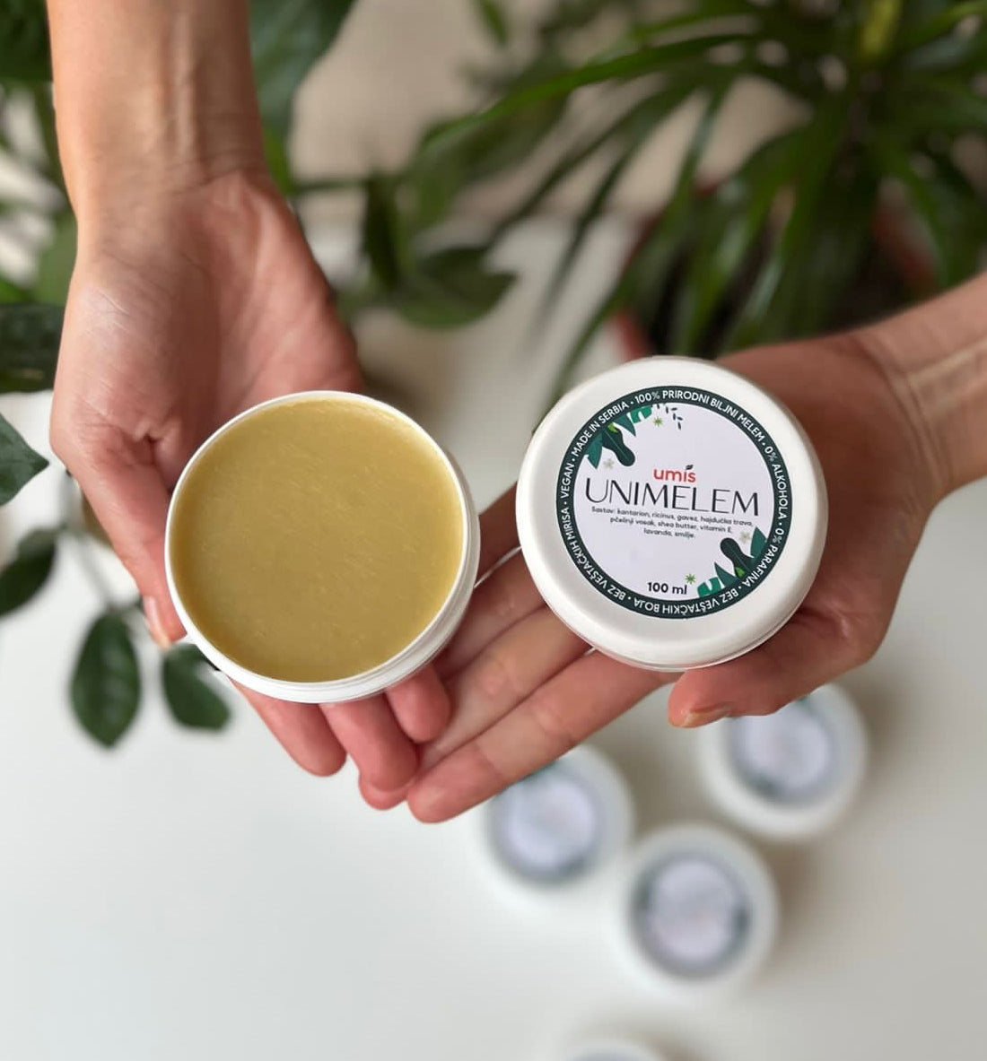 Ruke koje drže otvorenu kutiju melema "Unimelem." Jedna ruka drži poklopac na kojem se vidi naziv i informacije o sastavu proizvoda, dok druga ruka drži otvorenu kutiju s kremastim sadržajem žute boje melema. U pozadini se nalaze još nekoliko zatvorenih pakovanja melema za psorijazu i opekotine