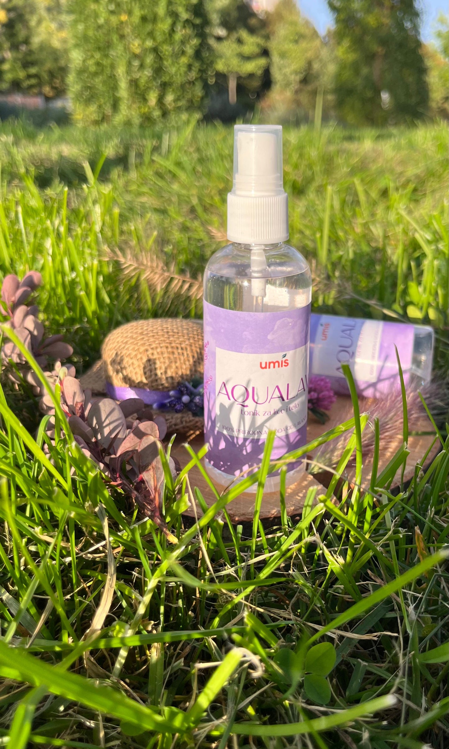 Bočica AQUALAV tonika za lice i telo, smeštena u travnatom okruženju, uz šešir od prirodnih materijala i ljubičasto cveće, što naglašava prirodnost i estetsku privlačnost proizvoda, dok sunčeva svetlost stvara osvežavajuću atmosferu. Tonik, napravljen od 100% prirodne lavandine vode, pomaže u borbi protiv akni, smanjuje upale i crvenilo, hidrira kožu, vraća njen prirodni pH balans i štiti je od štetnih spoljašnjih uticaja, čineći ga idealnim za svakodnevnu negu i održavanje zdravog tena.
